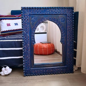 May include: A blue wooden mirror with an ornate carved frame and an arched opening. The mirror is reflecting a room with a pouf and a laptop.