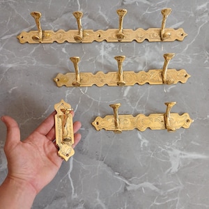 Puede incluir: Varios colgadores de abrigo antiguos de latón con diseños florales ornamentados. Los colgadores tienen múltiples ganchos y están montados sobre una superficie gris con patrón de mármol. Una mano sostiene un solo gancho.