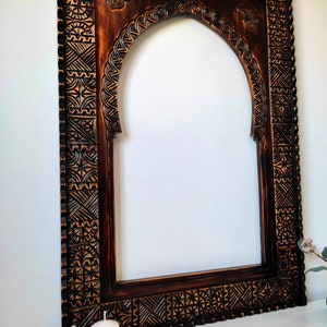 May include: A dark brown wooden mirror frame with intricate carvings. The frame has an arched top and a rectangular shape. The carvings are in a geometric pattern.
