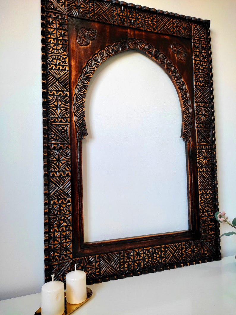May include: A dark brown wooden mirror frame with intricate carvings. The frame has an arched top and a rectangular shape. The carvings are in a geometric pattern.