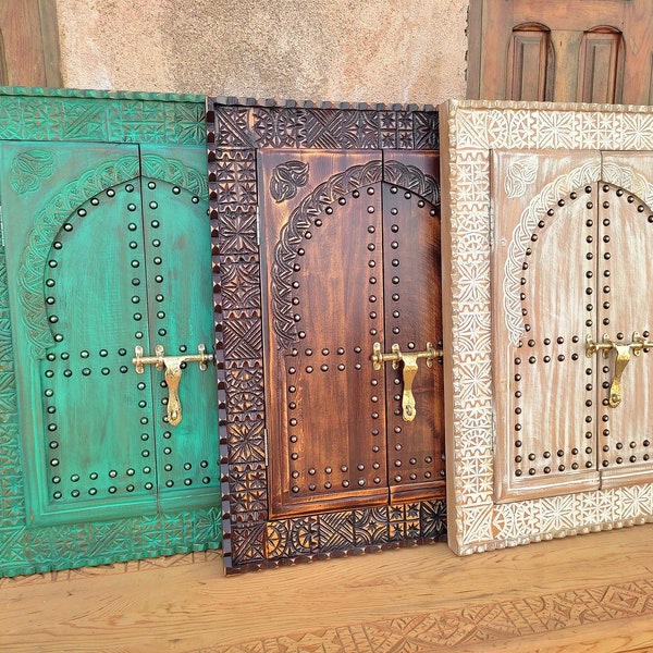 May include: Three rectangular wooden wall decor pieces shaped like arched doors. Each features intricate carved borders, a central arched panel with metal stud accents, and a brass-coloured sliding bolt latch. The pieces are finished in green, dark brown, and white.