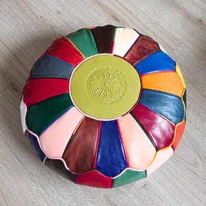 Peut inclure: Un pouf rond en cuir multicolore avec un motif géométrique. Le pouf présente un cercle central vert olive avec un motif en relief. Les panneaux environnants sont de différentes couleurs, dont le rouge, le bleu et le marron.