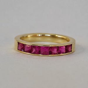 Puede incluir: Un anillo de oro con un engaste de canal que presenta siete piedras preciosas de corte cuadrado en un tono rojo rosado vibrante, dispuestas en fila a lo largo de la mitad frontal. El metal tiene un acabado pulido y las piedras están al ras.