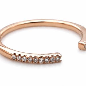 May include: Rose gold open ring with a row of small, clear gemstones. The ring has a modern, minimalist design with an open front. The gemstones are evenly spaced along the band, adding a touch of sparkle.