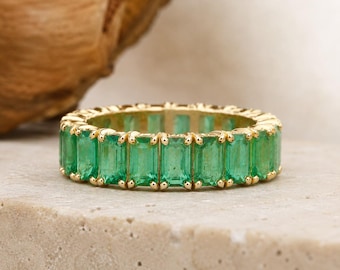 May include: A gold eternity band featuring a continuous row of vibrant green emerald-cut gemstones. Each rectangular emerald is set in a polished gold prong setting, creating a uniform and circular design that encircles the entire finger.
