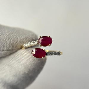 May include: A gold ring featuring two oval-cut, deep red gemstones. The ring's band is split, with one side adorned with small, clear stones and the other with dark stones. The ring is held against a neutral background.