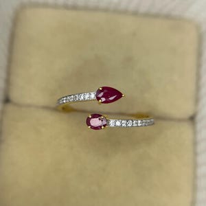May include: A gold-toned ring featuring two pear-shaped, deep red gemstones. The ring band is split, with each side embellished with small, clear gemstones. The ring is set against a neutral background.