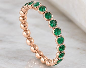 May include: A rose gold eternity ring featuring a series of round, vibrant green gemstones set in individual bezels. The band is constructed from a sequence of polished metal spheres that alternate with the circular green stones throughout.