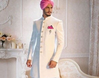 Pode incluir: Um terno branco de manga comprida com gola mandarim, combinado com calças brancas. A roupa apresenta um lenço de bolso rosa e um turbante rosa a condizer. O conjunto é completado com sapatos sociais castanhos, criando um visual formal e elegante.