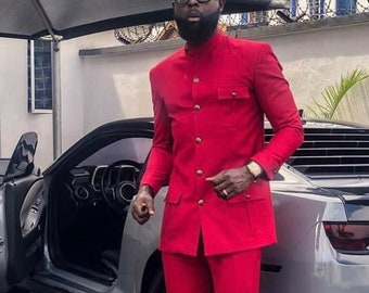 May include: A man wearing a red suit with a mandarin collar and gold buttons. He is standing in front of a gray car and wearing white sneakers.