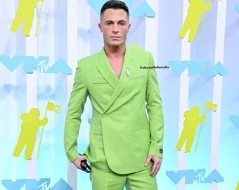 May include: A man wearing a bright green suit with a double-breasted jacket and wide-leg pants. He is standing on a red carpet with a backdrop of white and blue panels featuring the MTV logo.