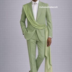 May include: A light green suit with a draped fabric detail. The suit includes a blazer, trousers, and a white dress shirt. The model is wearing a decorative brooch. The suit is a solid color and has a modern design.