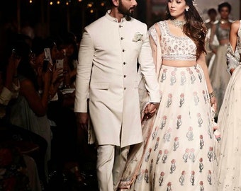 Pode incluir: Um casal com roupas tradicionais indianas brancas. A mulher está usando uma saia branca com estampa floral e um top branco. O homem está usando uma camisa branca de manga comprida e calças brancas. Eles estão andando de mãos dadas em uma passarela.