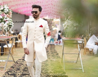 May include: A man wearing a white sherwani with gold buttons and a red pocket square. He is wearing white trousers and brown shoes. He is standing on a black and gold patterned rug.