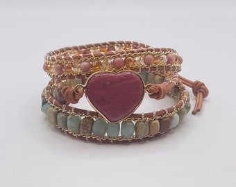 May include: A multi-layered wrap bracelet with a heart-shaped pink stone charm. The bracelet is made with brown leather, gold chain, and various colored beads.