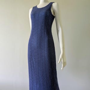 May include: A long, blue dress on a mannequin. The dress is sleeveless and reaches the floor, featuring a textured pattern and a slight shimmer. The hem has a patterned trim. The dress is set against a plain white backdrop.