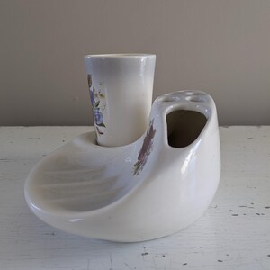 May include: White ceramic toothbrush holder with a floral design. The holder has a separate compartment for a toothbrush and a small cup for toothpaste.