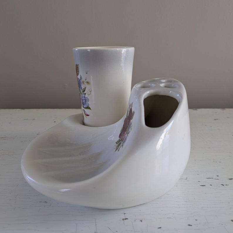 May include: White ceramic toothbrush holder with a floral design. The holder has a separate compartment for a toothbrush and a small cup for toothpaste.