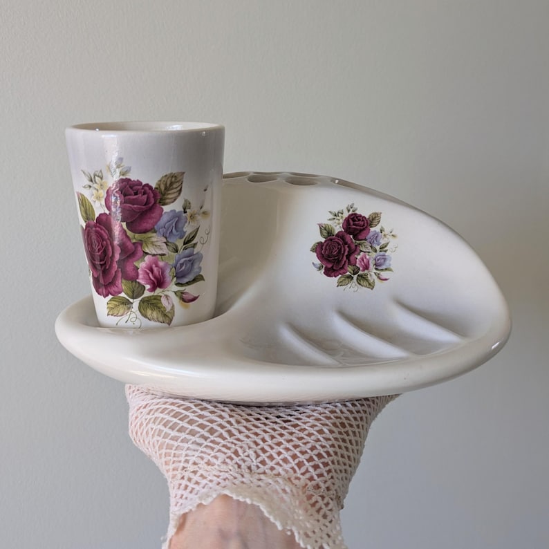 May include: White ceramic bathroom set with a toothbrush holder and a cup. The set features a floral design with pink and purple roses.