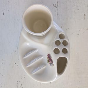 May include: White ceramic toothbrush holder with a floral design. It has a cup for toothpaste, a soap dish, and multiple slots for toothbrushes.