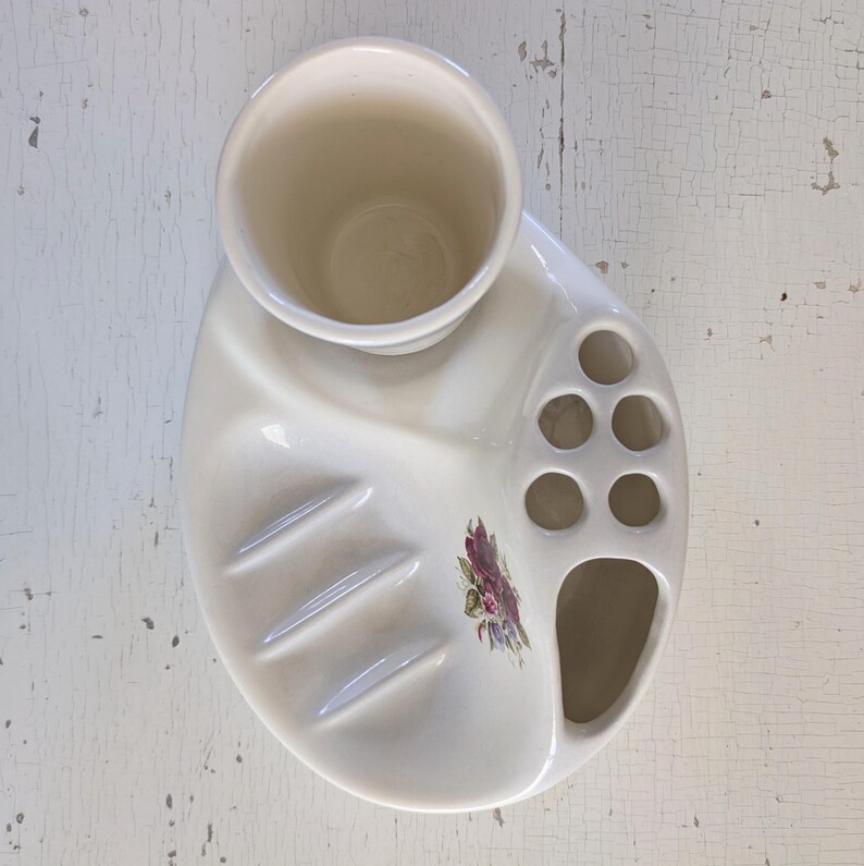 May include: White ceramic toothbrush holder with a floral design. It has a cup for toothpaste, a soap dish, and multiple slots for toothbrushes.