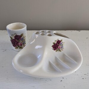 May include: White ceramic bathroom set with a toothbrush holder, soap dish, and tumbler. The set features a floral design with pink roses.