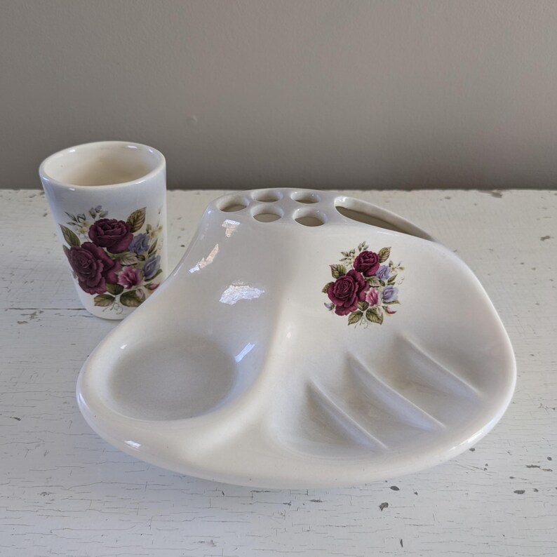 May include: White ceramic bathroom set with a toothbrush holder, soap dish, and tumbler. The set features a floral design with pink roses.