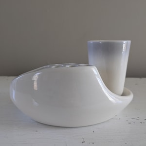 May include: White ceramic flower vase with a separate white ceramic cup. The vase has a unique, curved shape and multiple holes for flower stems. The cup has a light gray ombre effect.
