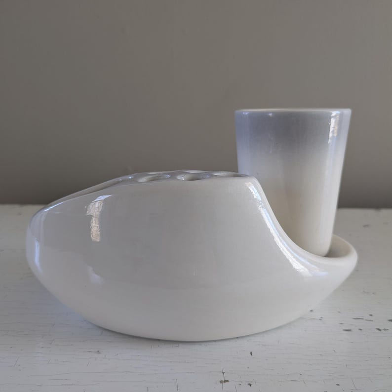 May include: White ceramic flower vase with a separate white ceramic cup. The vase has a unique, curved shape and multiple holes for flower stems. The cup has a light gray ombre effect.