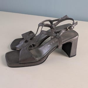 Pode incluir: Um par de sandálias Nine West cinza metálico com salto. As sandálias têm bico quadrado, design em T e fecho de fivela. Os saltos têm aproximadamente 7,6 cm de altura.
