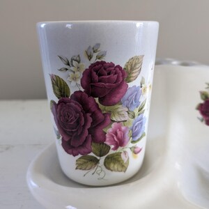 May include: White ceramic toothbrush holder with a floral design. The holder has three cups, one of which is shown in the image. The cup has a white background with a floral design featuring burgundy roses, pink and blue flowers, and green leaves.