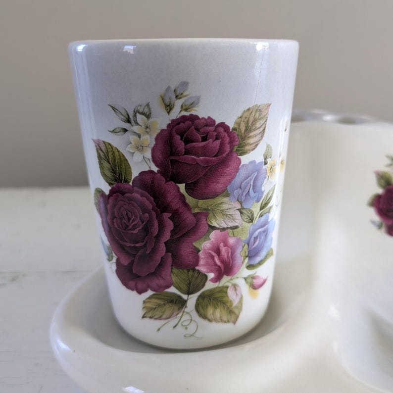 May include: White ceramic toothbrush holder with a floral design. The holder has three cups, one of which is shown in the image. The cup has a white background with a floral design featuring burgundy roses, pink and blue flowers, and green leaves.