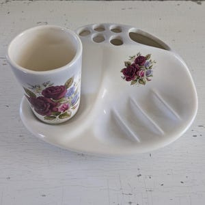 May include: White ceramic bathroom set with a toothbrush holder, soap dish, and tumbler. The set features a floral design with pink roses and green leaves.