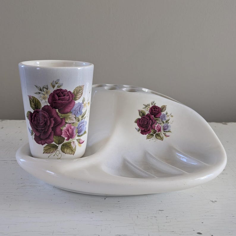 May include: White ceramic bathroom set with a toothbrush holder and a tumbler. Both pieces feature a floral design with pink and purple roses.