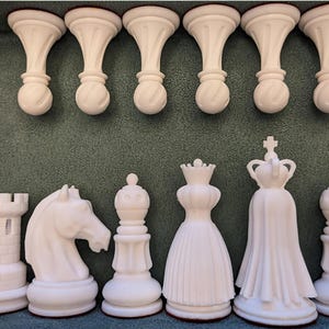 May include: A set of white chess pieces including a rook, knight, bishop, queen, king, and pawns. The pieces feature smooth, matte surfaces with detailed carvings on the knight and crown, arranged in rows against a dark green textured surface.