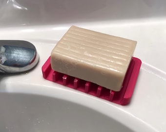 Può includere: Una saponetta rettangolare beige è appoggiata su un portasapone rosa acceso. Il sapone ha una superficie a coste. Il portasapone in plastica ha un design a fessure. Gli oggetti sono su una superficie bianca.