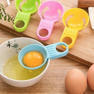 Puede incluir: Cuatro separadores de huevos de plástico de colores brillantes con un mango y un tazón pequeño para separar las yemas de las claras. Los separadores son verde, rosa, amarillo y azul. Un separador se muestra separando una yema de huevo en un tazón blanco.