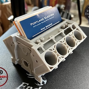 May include: A light gray 3D printed business card holder shaped like a V8 engine block. The holder features four visible cylinder bores and holds a stack of blue business cards with the text Portland Electric Appliance Sales With Service Mike Bibiskie.