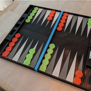 May include: A black backgammon board with red and green counters. The board features a blue stripe and dice. The game is set on a light-coloured surface.