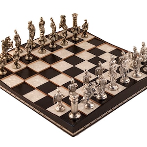 Puede incluir: Un juego de ajedrez con piezas de plata y oro sobre un tablero de ajedrez blanco y negro. El tablero está hecho de madera y tiene un borde decorativo.