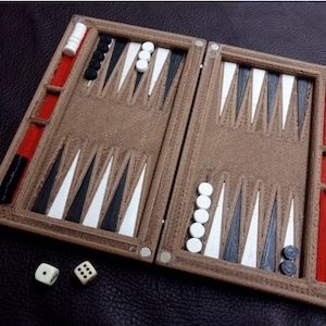 May include: A brown backgammon board with black, white, and red detailing. The board is open, showing the playing surface with triangular points. Dice and game pieces are present, ready for play. The game board is on a dark surface.