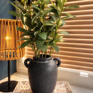 May include: A dark charcoal grey ceramic vase with two side handles containing a faux olive branch with green leaves and dark olives. The vase sits on a rectangular white tray with a brown leaf pattern and the text D&R Botanical Blooms & Homeware.