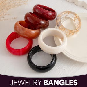 Könnte beinhalten: Eine Auswahl an farbenfrohen Armreifen in Rot-, Braun-, Weiß-, Schwarz- und transparenten Farbtönen mit Goldblatt-Akzenten. Die Schmuckarmreifen sind aus Kunstharz gefertigt und auf einer weißen Oberfläche präsentiert.