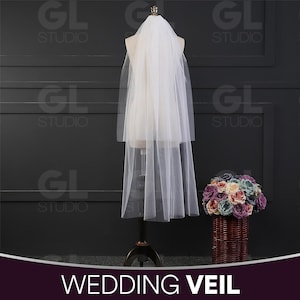 Peut inclure: Un voile de mariage blanc à deux niveaux, présenté sur un mannequin. Le voile est transparent et délicat, avec un aspect doux et fluide. Un bouquet de fleurs colorées dans un panier en osier est à côté. Le texte "WEDDING VEIL" est en bas.