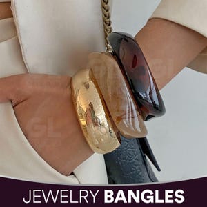 Op de afbeelding: Vier armbanden in verschillende kleuren en texturen. De armbanden omvatten een getextureerd goud, een doorschijnend beige, een schildpadbruin en een effen donkerbruin. De tekst "JEWELRY BANGLES" is onderaan de afbeelding te zien.