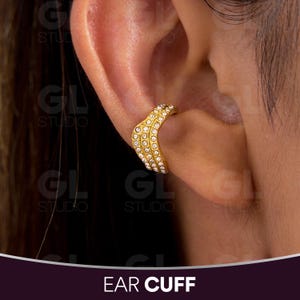 May include: Gold-toned ear cuff with a chevron design, adorned with small, sparkling crystals. The ear cuff is shown on an ear, with the text "EAR CUFF" at the bottom.