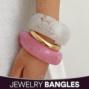 Op de afbeelding: Een stapel van drie armbanden. Een dikke, doorschijnende witte armband met een marmereffect ligt bovenop een roze gemarmerde armband. Een goudkleurige metalen armband zit ertussen. De tekst "JEWELRY BANGLES" staat onderaan.