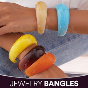 Könnte beinhalten: Eine Auswahl an auffälligen, geschwungenen Armreifen in Gelb, Bernstein, Orange, Braun und Blau. Der Schmuck wird an Handgelenken vor einem Jeanshintergrund gezeigt. Der Text "JEWELRY BANGLES" befindet sich am unteren Bildrand.