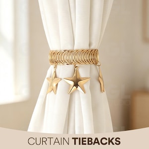 May include: A gold-toned metal curtain tieback featuring a series of interconnected circular rings forming a wide band. Three smooth, five-pointed star charms in a matching gold finish hang from the bottom edge of the band, securing white fabric.