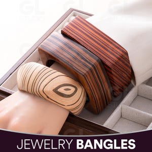 Può includere: Braccialetti in legno in varie tonalità di marrone, esposti in un portagioie. Un braccialetto è di colore chiaro con un motivo a venature circolari, mentre gli altri sono più scuri con motivi a venature lineari. Il testo "JEWELRY BANGLES" è in basso.